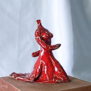Puede incluir: Escultura de cerámica roja de una bailarina con un vestido fluido. La figura está en movimiento con los brazos extendidos. La escultura tiene un acabado brillante con detalles blancos. La base es un bloque de madera. El texto "DANCE IS" es visible.