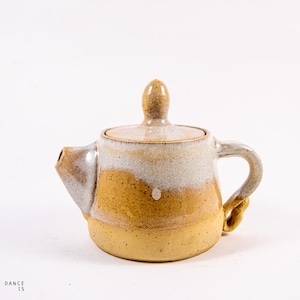 Può includere: Una teiera in ceramica fatta a mano con una glassa macchiettata in tonalità di marrone e bianco. La teiera ha un corpo arrotondato e un coperchio con un pomo.