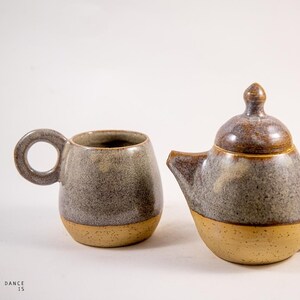Può includere: Una tazza in ceramica con manico circolare e una teiera abbinata con coperchio, entrambe caratterizzate da uno smalto bicolore con parte superiore grigia maculata e base beige solida. Il testo DANCE IS è stampato in basso a sinistra.