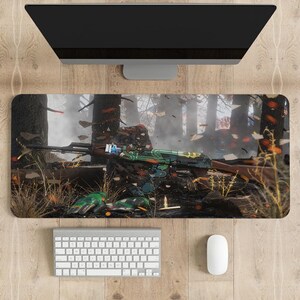 May include: A rectangular desk mat featuring a graphic print of a green and black rifle resting on a dark forest floor. The design includes scattered leaves, a single bullet, and a pair of green gloves, set against a backdrop of trees.