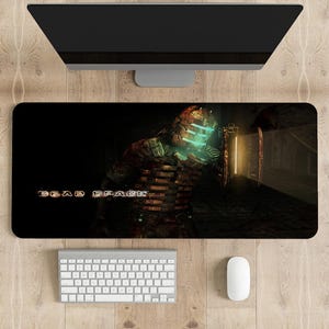 May include: A black gaming mouse pad with a dark background featuring a character from the video game Dead Space. The character is wearing a dark brown and green spacesuit with a glowing blue helmet. The text "Dead Space" is printed in white on the mouse pad.