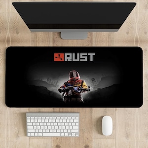 Puede incluir: Alfombrilla de ratón rectangular negra con el título del juego "RUST" en blanco y rojo. El diseño incluye una figura con un atuendo colorido sosteniendo un arma, sobre un fondo oscuro y brumoso. Un teclado y un ratón blancos son visibles debajo.