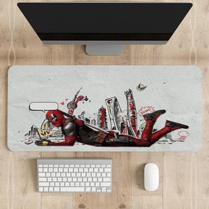May include: A rectangular desk mat featuring a comic-style illustration of Deadpool in a red and black suit, with a cityscape background. The mat includes a keyboard and mouse.