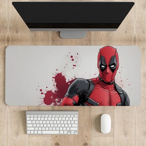 May include: A rectangular white desk mat featuring a graphic of Deadpool in his red and black costume, with red splatter accents. A computer keyboard and mouse are below the mat, and a computer monitor is above.