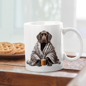 May include: White ceramic mug featuring a brown dog wrapped in a gray blanket, holding a cup of coffee. The dog illustration is detailed and realistic. The mug has a curved handle and is set on a wooden surface.