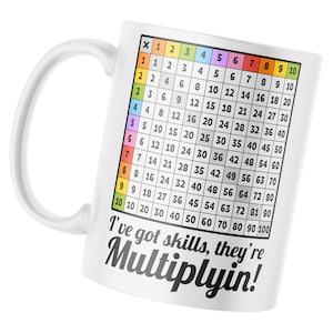 Pode incluir: Caneca de cerâmica branca com uma tabela de multiplicação impressa nela. A tabela vai de 1 a 10. A caneca tem uma alça preta e o texto "I've got skills, they're Multiplyin!" impresso na parte inferior.