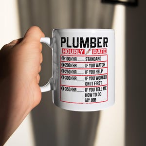 Puede incluir: Taza de cerámica blanca con la palabra "PLUMBER" en la parte superior y una lista de tarifas por hora. Las tarifas son para diferentes niveles de participación, desde estándar hasta decirle al fontanero cómo hacer su trabajo.