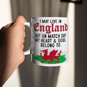 Può includere: Tazza in ceramica bianca con un drago rosso e una bandiera verde e bianca. Il testo sulla tazza recita "I may live in England but on match day my heart & soul belong to".