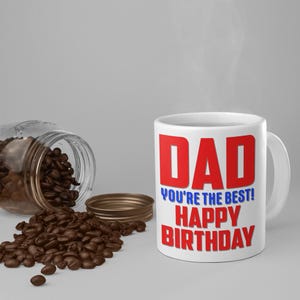 Può includere: Tazza da caffè in ceramica bianca con il testo "DAD YOU'RE THE BEST! HAPPY BIRTHDAY" in lettere rosse e blu. La tazza è appoggiata su una superficie grigia con chicchi di caffè sparsi intorno.
