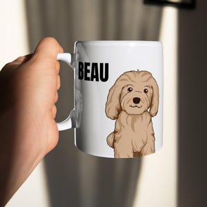 May include: White ceramic mug featuring the name "BEAU" in black block letters and a cartoon illustration of a tan-colored dog. The dog has floppy ears and a dark brown nose. The mug has a white handle.