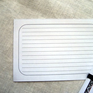 May include: White lined recipe cards with rounded corners. A stack of cards is visible, held together with a black elastic band. The cards have a decorative border and text. The background is a neutral linen fabric.