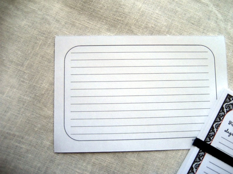 May include: White lined recipe cards with rounded corners. A stack of cards is visible, held together with a black elastic band. The cards have a decorative border and text. The background is a neutral linen fabric.