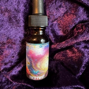 May include: A small, dark glass bottle with a black dropper top. The label features the text "Cottonwood Conjure" and "SWIFTWAY" over a colorful, swirling galaxy design. The bottle sits on a plush, purple velvet surface.