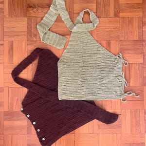 May include: Two handmade crochet tops in the image. One is a light olive green halter top with tie closures. The other is a deep burgundy wrap top with button closures. Both tops are made of a textured yarn and are displayed on a wooden surface.