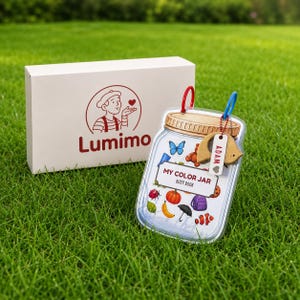 Könnte beinhalten: Eine weiße Schachtel mit dem Lumimo-Logo neben einem durchsichtigen, gläserförmigen Beschäftigungsbuch. Das Buch zeigt farbenfrohe Illustrationen eines Schmetterlings, eines Teddybären, eines Kürbisses und anderer Objekte. Das Buch trägt die Aufschrift "My Color Jar Busy Book".