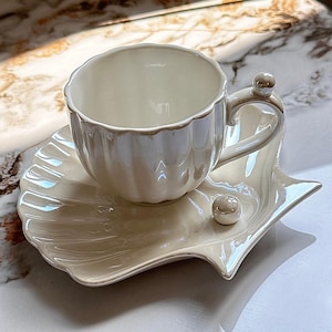 Può includere: Un set di tazza e piattino da tè color crema. La tazza ha un bordo smerlato e un manico curvo con un piccolo accento a sfera. Il piattino è a forma di conchiglia, con un accento a sfera abbinato. Il set ha una finitura perlata.