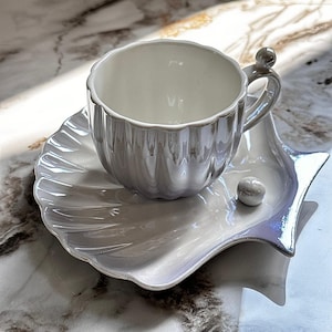 Può includere: Un set di tazza e piattino da tè bianco perlato. La tazza ha un design scanalato e un piccolo dettaglio di perla sul manico. Il piattino ha la forma di una conchiglia, con un accento di perla. Il set è in ceramica.