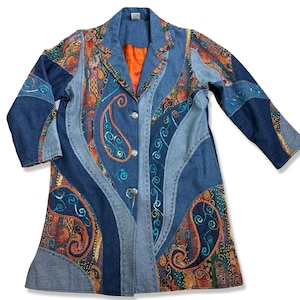 Puede incluir: Un abrigo largo de mezclilla con un diseño de patchwork. El abrigo presenta una variedad de tonos de mezclilla azul, con acentos de tela estampada de cachemira y floral en naranja, rojo y verde azulado. El abrigo tiene tres botones plateados y un forro naranja.