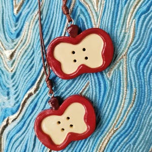 May include: Two red apple-shaped pendants with a cream-coloured centre and four black buttons. The pendants are hanging from a brown cord.