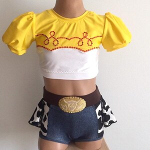 May include: A yellow and white crop top with a red and white trim and a cow print skirt with a brown belt and a gold buckle. The belt buckle has a cow head design.