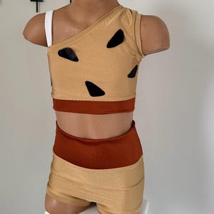 May include: A two-piece dance costume with a brown top and matching shorts. The top has a one-shoulder design and features black triangle shapes. The shorts have a brown waistband.