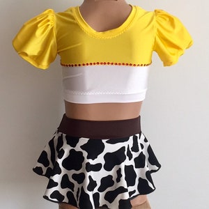 May include: A yellow and white crop top with a brown and black cow print skirt. The top has red rhinestones around the neckline and the skirt has a brown waistband.