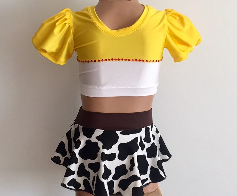 May include: A yellow and white crop top with a brown and black cow print skirt. The top has red rhinestones around the neckline and the skirt has a brown waistband.