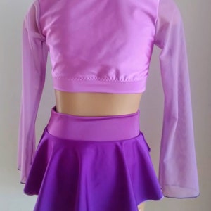 Op de afbeelding: Een tweedelig danskostuum in paarse tinten. De top is een lichtpaarse crop top met doorschijnende mouwen. De rok is donkerpaars, uitlopend, met een brede tailleband. Het kostuum wordt op een mannequin gepresenteerd.