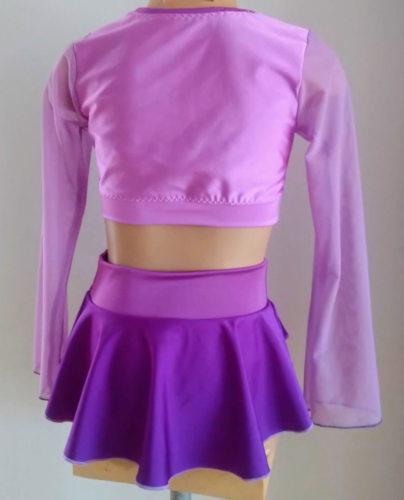 Op de afbeelding: Een tweedelig danskostuum in paarse tinten. De top is een lichtpaarse crop top met doorschijnende mouwen. De rok is donkerpaars, uitlopend, met een brede tailleband. Het kostuum wordt op een mannequin gepresenteerd.
