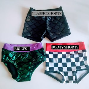 Op de afbeelding: Drie korte broeken worden getoond. De bovenste is zwart met een schubbenpatroon en de tekst "CLASSIC SHORTS". De middelste is groen met een schubbenpatroon en een paarse tailleband, met het label "BRIEFS". De onderste is geruit met een rode tailleband, met het label "BOOTY SHORTS".