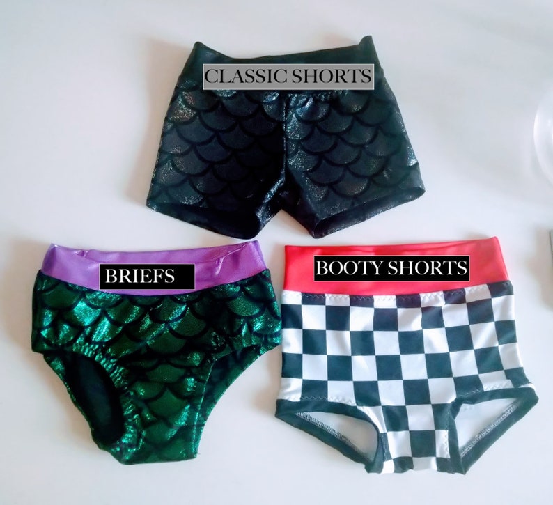 Op de afbeelding: Drie korte broeken worden getoond. De bovenste is zwart met een schubbenpatroon en de tekst "CLASSIC SHORTS". De middelste is groen met een schubbenpatroon en een paarse tailleband, met het label "BRIEFS". De onderste is geruit met een rode tailleband, met het label "BOOTY SHORTS".