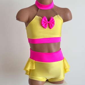 May include: A yellow and pink dance costume set. The set includes a halter top with a pink bow, a matching choker, and shorts with a ruffled hem. The costume has a bright yellow base with pink accents.