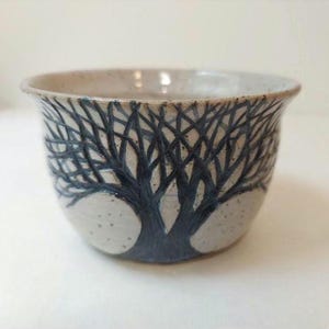May include: A ceramic bowl with a tree design. The bowl is a light beige color with a dark blue tree design wrapping around the exterior. The tree design is detailed with many branches.