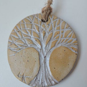 May include: A round, beige ceramic ornament with a white tree of life design. The ornament has a hole at the top for hanging and is attached to a brown twine string.