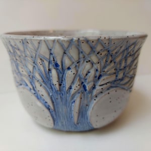 May include: A ceramic bowl with a tree design. The bowl is light gray with a blue tree design and black speckles. The tree branches are etched into the bowl's surface.