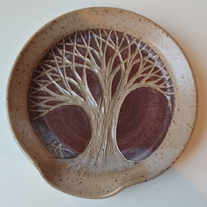 May include: A ceramic spoon rest with a brown and white glaze. The spoon rest features a raised design of a tree with branches.