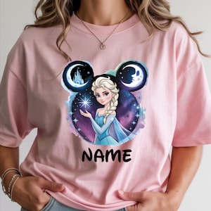 May include: Light pink t-shirt featuring a graphic of a princess with a blue dress and a braided hairstyle. The design includes a starry background, a castle, and the word "NAME" in black.