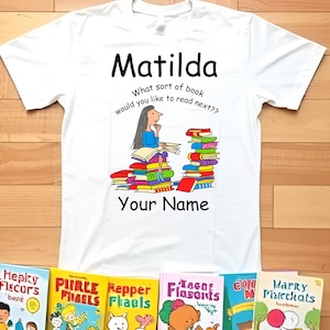 Puede incluir: Camiseta blanca con el nombre "Matilda" y una ilustración de una persona leyendo libros. El texto "What sort of book would you like to read next??" está encima de la ilustración, y "Your Name" está debajo.
