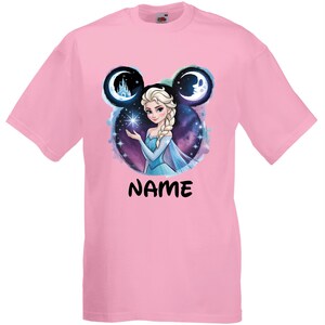 May include: Pink t-shirt featuring a graphic of a princess with a blue dress, holding a star. The design is set within a Mickey Mouse-shaped frame with a castle and moon. The word "NAME" is printed in black.