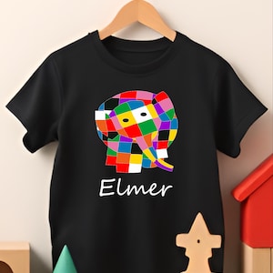 Puede incluir: Camiseta negra con un diseño de elefante de patchwork colorido y la palabra "Elmer" en escritura blanca. El diseño del elefante incluye cuadrados rojos, amarillos, verdes, azules y morados. La camiseta está colgada de una percha de madera.