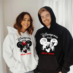 Może przedstawiać: Biała i czarna bluza z nadrukiem w kształcie uszu Myszki Miki. Nadruk zawiera sylwetkę zamku Kopciuszka, fajerwerki, Dzwoneczek i napis "Disneyland 2025". Pod nadrukiem znajduje się napis "Your Name".
