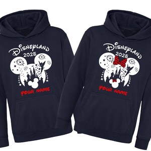 Może przedstawiać: Dwie granatowe bluzy z kapturem z białym nadrukiem uszu Myszki Miki z zamkiem i fajerwerkami w środku. Tekst "Disneyland 2025" znajduje się nad uszami, a "Your Name" pod uszami.