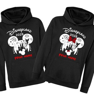 Może przedstawiać: Dwie czarne bluzy z kapturem z białym nadrukiem uszu Myszki Miki z tekstem "Disneyland 2025" i "Twoje Imię" nadrukowanym z przodu.