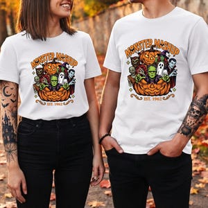 May include: A white t-shirt featuring a colorful graphic of various monsters, including Frankenstein's monster, a ghost, and a vampire, emerging from a carved jack-o'-lantern. The text reads "MONSTER MASHUP" at the top and "EST. 1962" at the bottom.