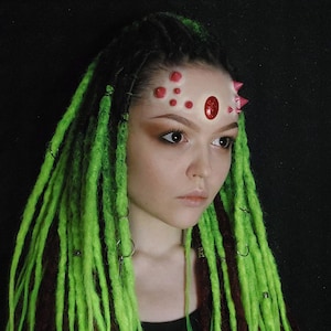 May include: A person with green and brown dreadlocks, wearing a red and pink headpiece. The headpiece features a large oval gem and several smaller, round, and spiky accents. The person has dark eye makeup and a neutral lip colour.