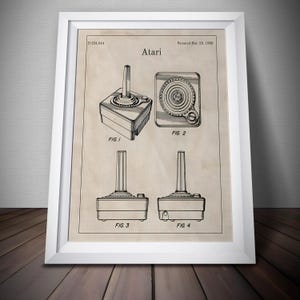 Puede incluir: Impresión enmarcada de patente Atari vintage. La ilustración en blanco y negro muestra diferentes vistas del joystick Atari. La impresión incluye el logotipo de Atari, información de patente y diagramas de figuras. El marco es blanco y el fondo es beige neutro.