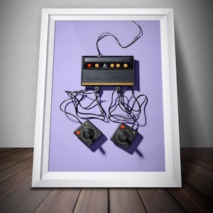 May include: Framed print featuring a vintage Atari game console and two joysticks against a purple background. The console is black and gold with red and yellow buttons. The joysticks are black with red buttons and circular control sticks.
