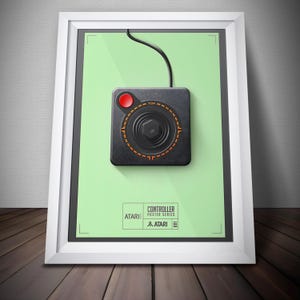 May include: Framed poster showing a black Atari joystick controller with a red button and orange detailing on a light green background. The poster includes the Atari logo and the text "Controller Poster Series."