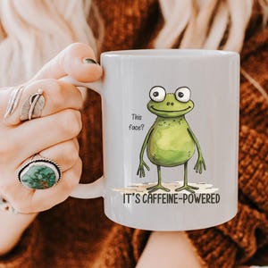 以下が含まれることがあります： 漫画のカエルのイラストと「This face?」と「IT'S CAFFEINE-POWERED」の文字が入った、明るい灰色のセラミックマグカップ。カエルは緑色で、大きな目と笑顔の顔をしています。マグカップには白い持ち手が付いています。