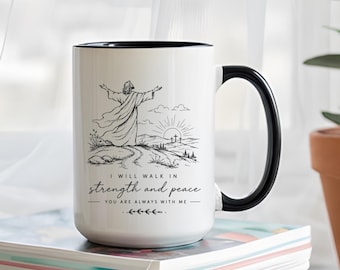 May include: A white ceramic mug with a black handle and interior. The front features a black line art illustration of a figure with arms outstretched toward a sunrise and three crosses. Text reads: I WILL WALK IN strength and peace YOU ARE ALWAYS WITH ME.